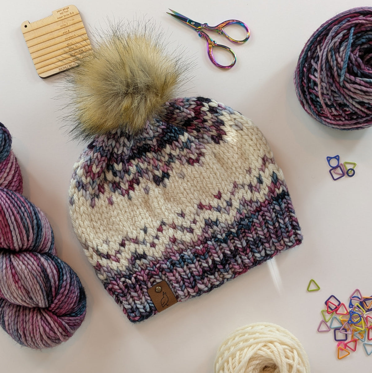 A hand-knit beanie with a large beige faux-fur pom-pom, featuring a cream, pink, and green variegated fair-isle pattern and a small brown suede quail logo patch. The beanie is styled in a flat-lay arrangement with yarn balls, scissors, and colorful stitch markers on a light beige background.