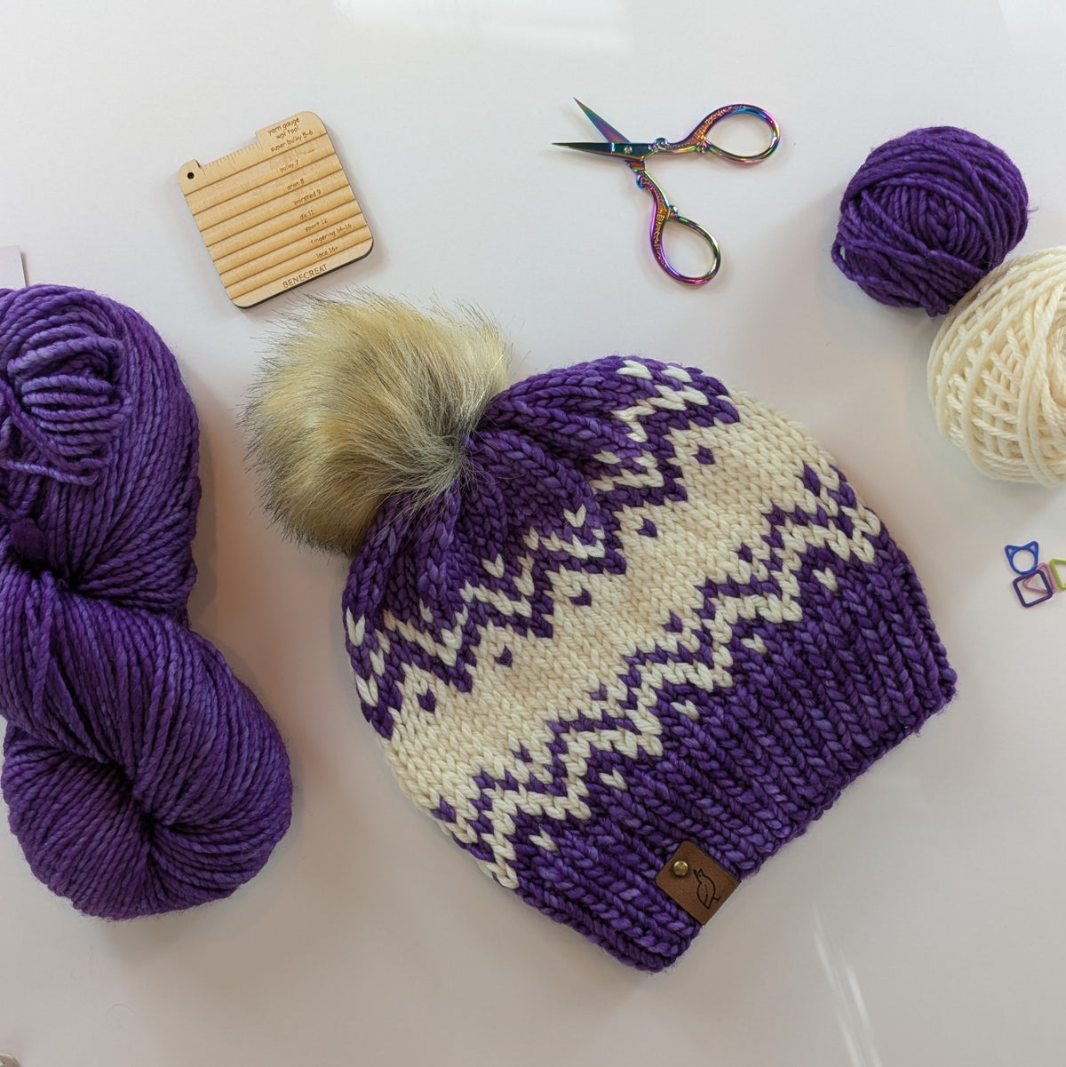 A purple and cream knit fair-isle beanie with a fluffy beige pom-pom, arranged on a white surface with yarn balls, a wooden gauge tool, and small scissors.