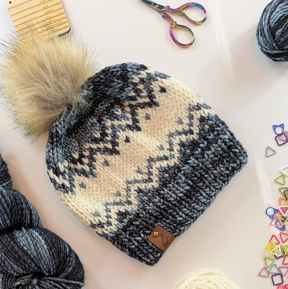 A knitted pom-pom beanie hat with a cream and dark gray and slate fair isle pattern, styled on a white surface with yarn, scissors, and colorful triangle stitch markers.