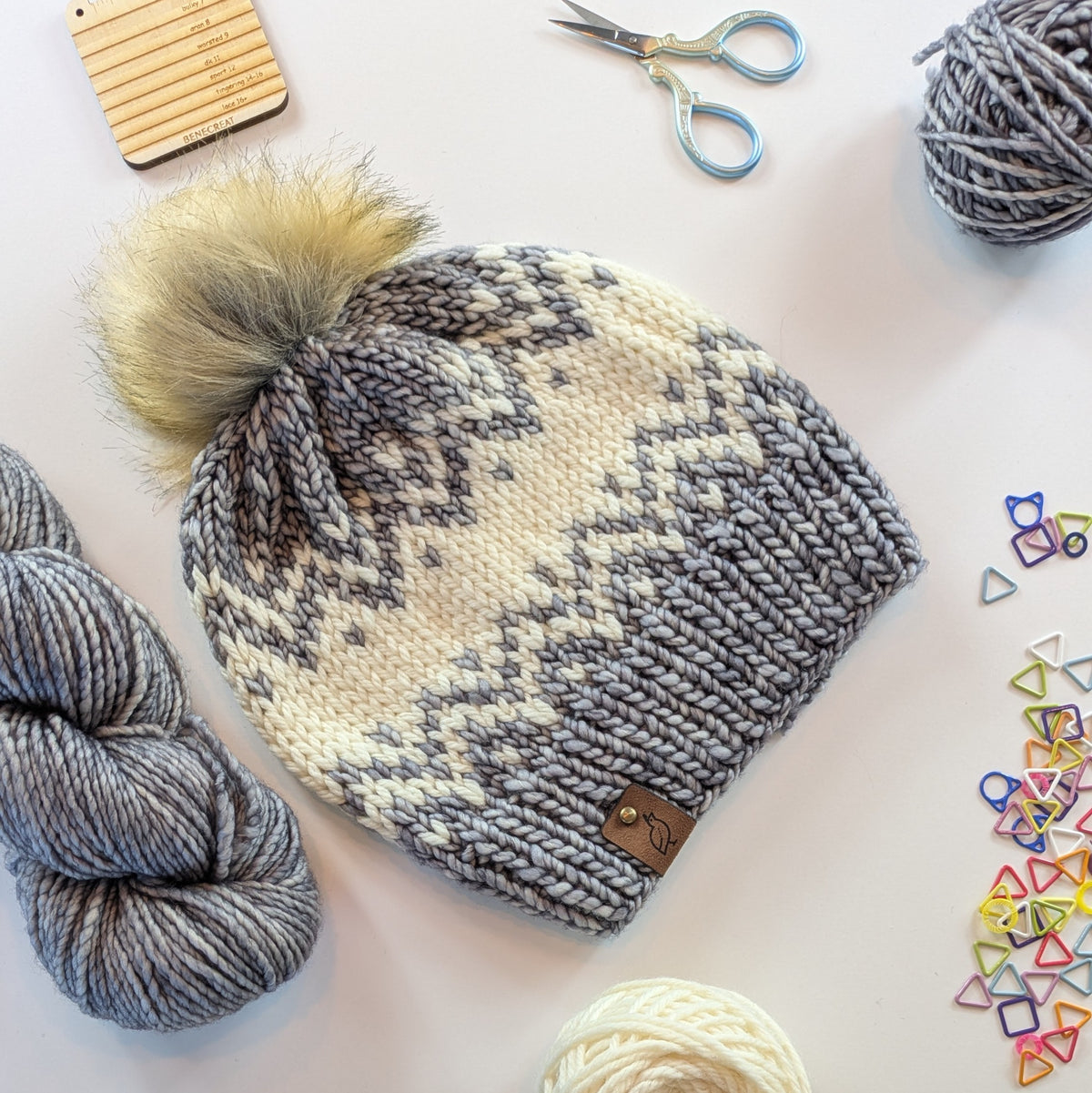 A hand-knit gray and cream beanie with a large beige pom-pom, featuring a Fair Isle style pattern. The beanie is styled in a flat-lay arrangement with knitting yarn, scissors, and colorful yarn markers on a white background.