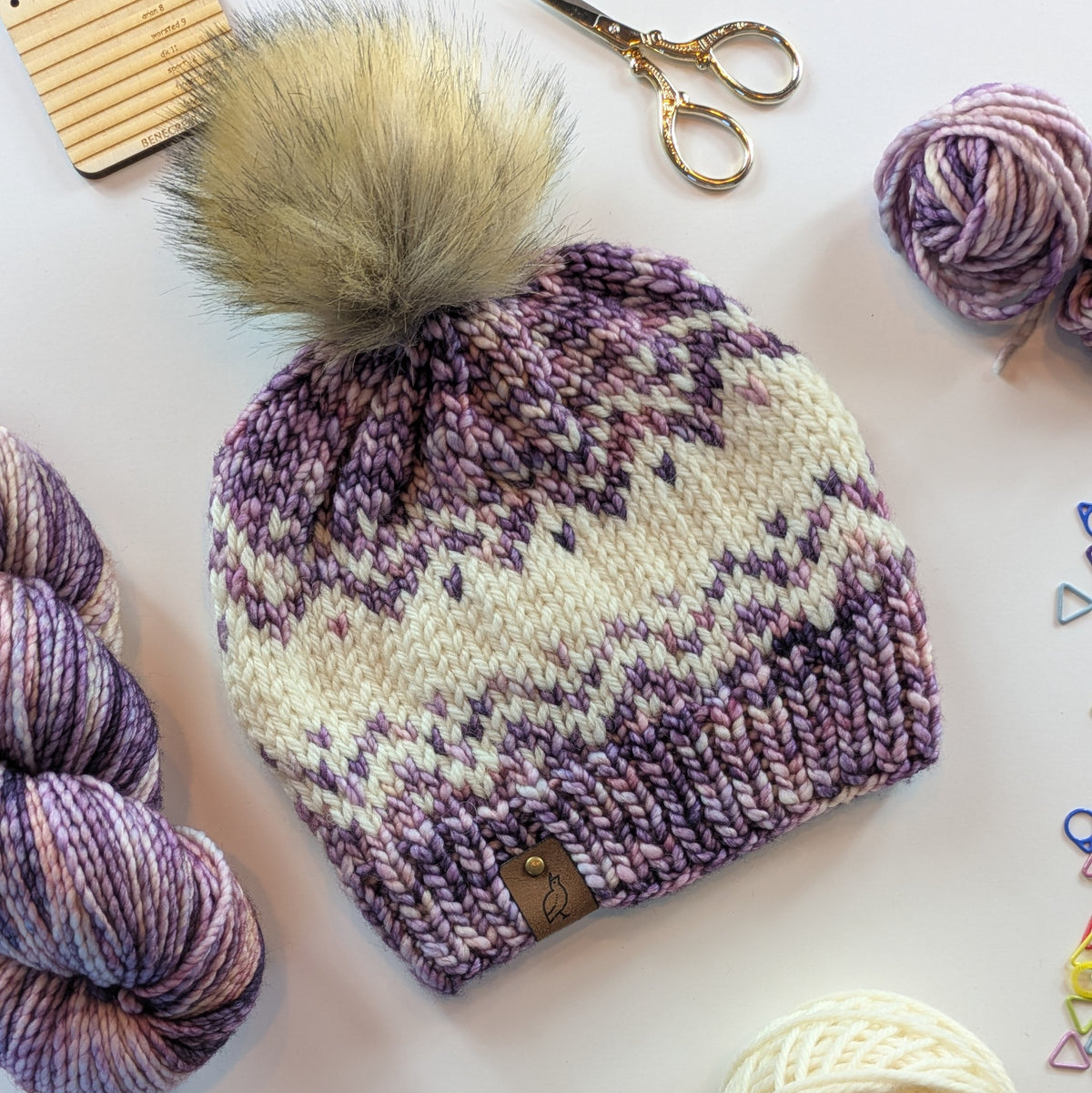 A hand-knit beanie hat with a large beige faux fur pom-pom, featuring a purple and cream Fair Isle pattern. The hat is styled on a white surface with knitting yarn, scissors, and a wooden yarn tool.
