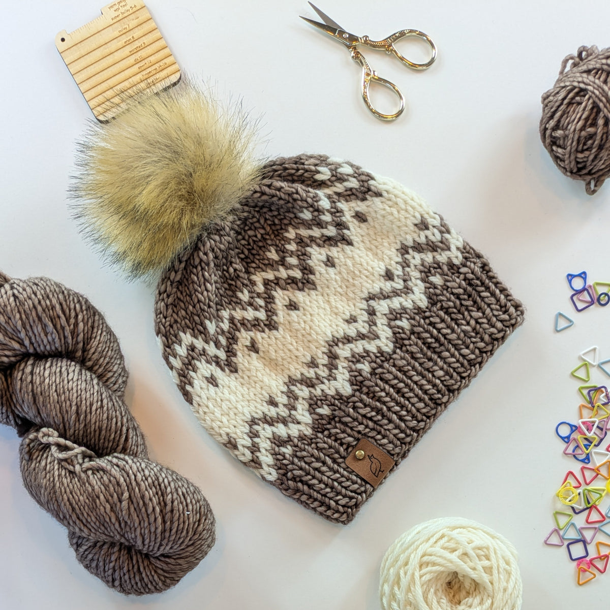 A hand-knit pom-pom beanie with a brown and cream fair isle pattern, styled in a flat lay with yarn, scissors, and knitting accessories on a white background.