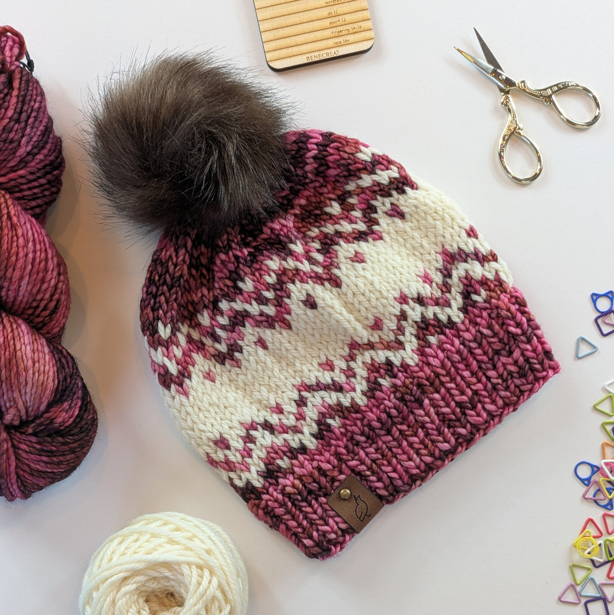 A hand-knit pom-pom beanie with a pink and cream Fair Isle pattern, styled on a white surface with yarn, scissors, and stitch markers.
