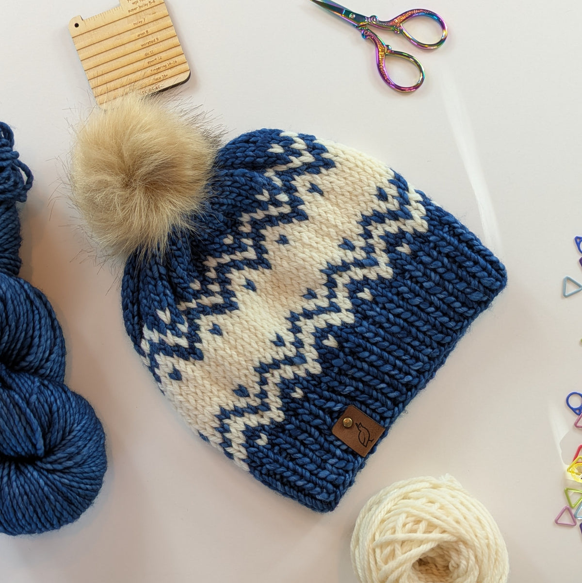 A hand-knit winter beanie with a cream and navy blue Fair Isle pattern, featuring a fluffy beige pom-pom on top and a small brown leather brand tag. The beanie is styled in a flat-lay arrangement with yarn balls, scissors, and knitting tools on a white background.