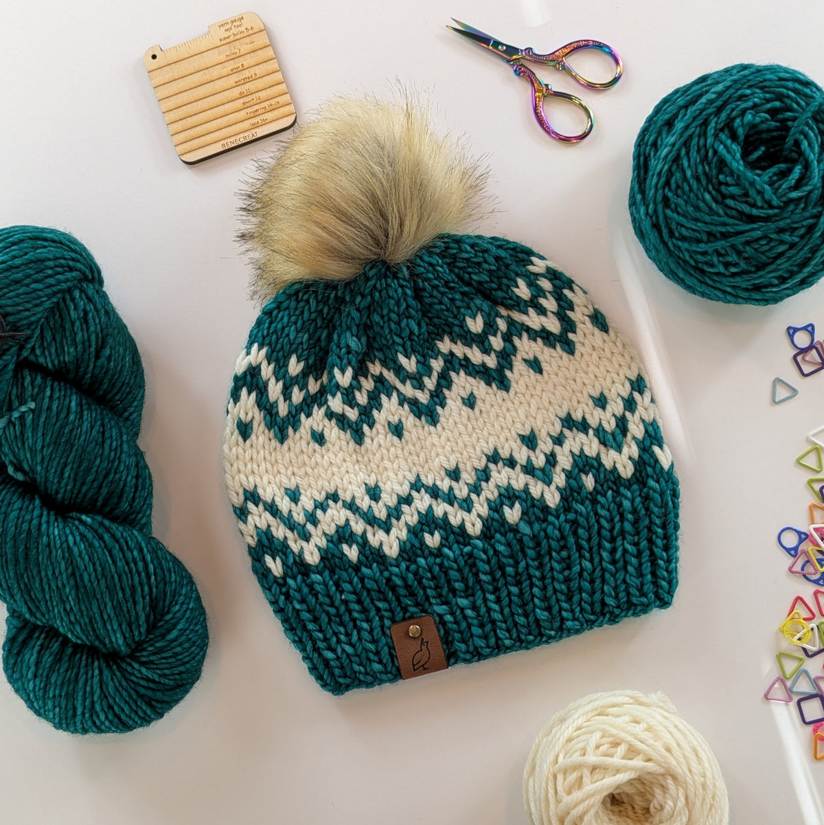 A knitted pom-pom beanie in teal and cream with a small brown leather logo tab, arranged on a white surface with yarn balls, scissors, and small colorful accessories around it.