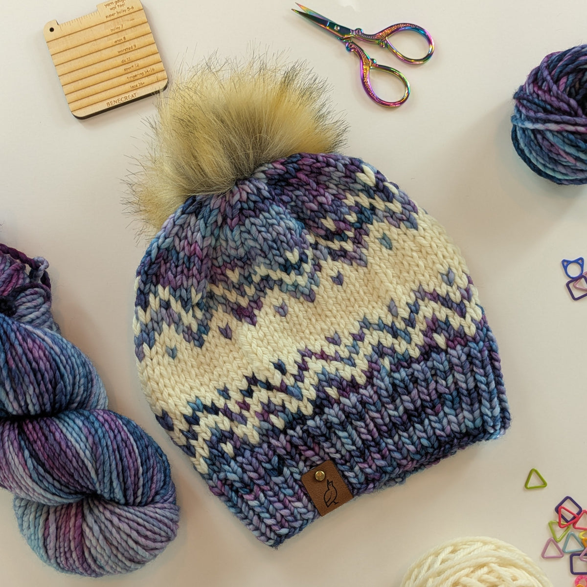 A hand-knit beanie with a fluffy faux-fur pom-pom, featuring a Fair Isle pattern in cream, purple, and blue-gray. The beanie is styled in a flat-lay arrangement with variegated yarn, rainbow scissors, and a wooden gauge tool on a light background.