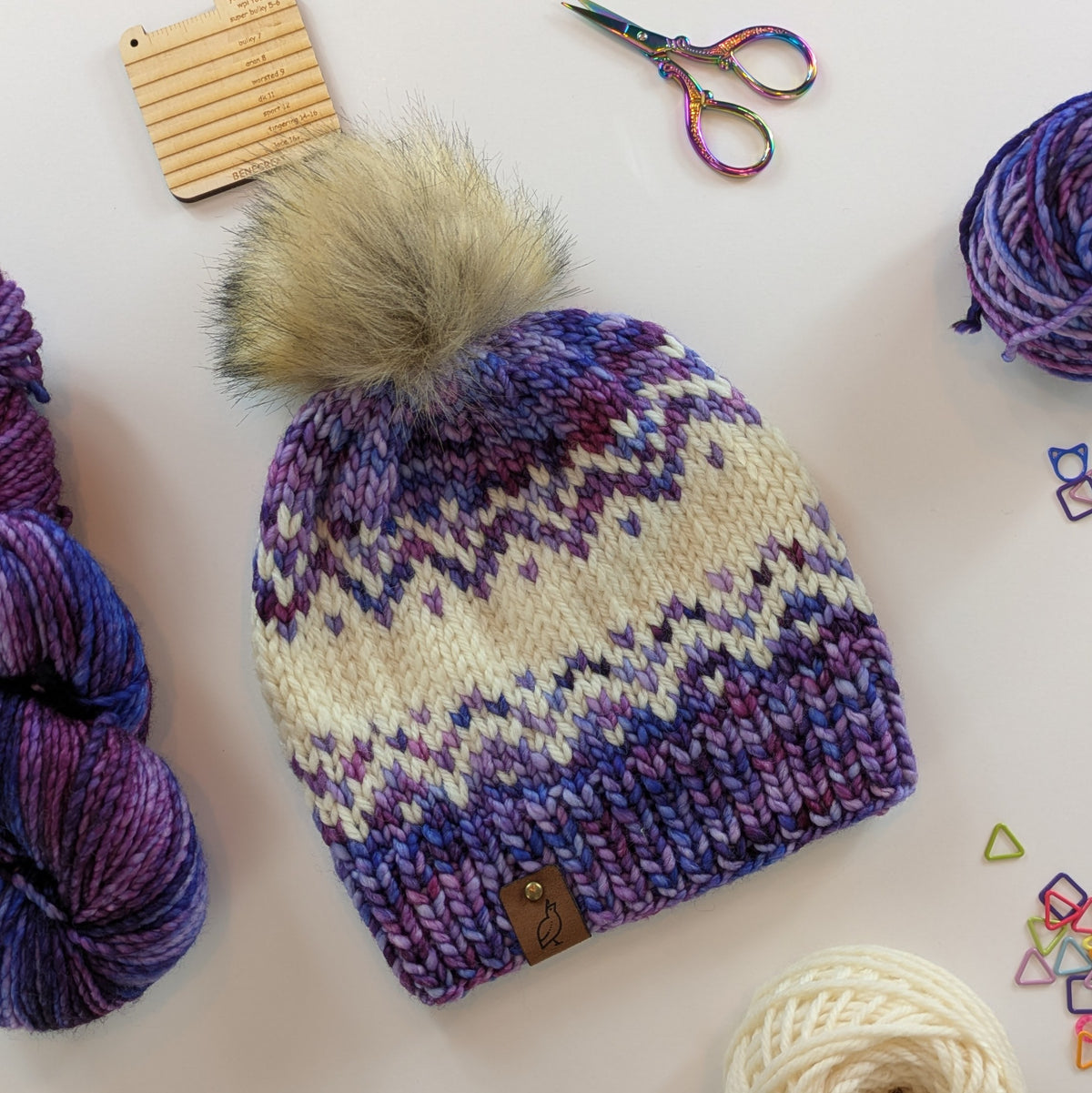 A hand-knit beanie hat with a large beige faux fur pom-pom, featuring a purple, cream, and blue Fair Isle pattern. The hat is styled in a flat-lay arrangement with yarn skeins, rainbow scissors, and small colorful geometric stitch markers on a white background.