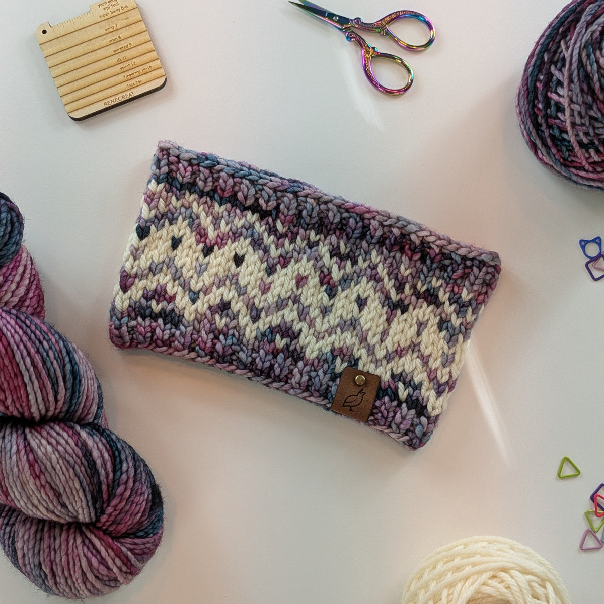 A hand knit fair isle ear warmer headband in a variegated pattern of cream with red, pink, and green yarn, styled in a flat-lay with knitting supplies on a white background.