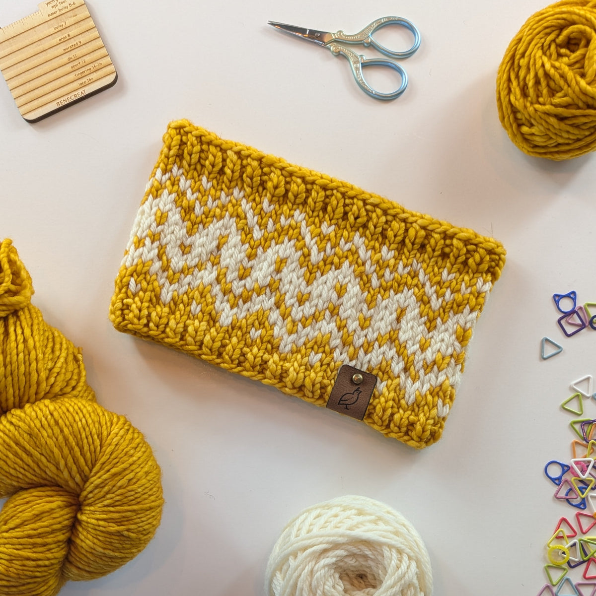 A hand knit fair isle ear warmer headband in golden yellow and cream, styled in a flat-lay with knitting supplies on a white background.