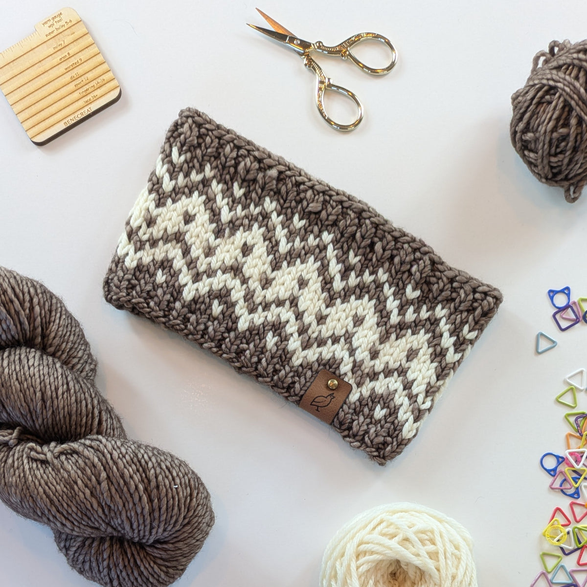 A hand knit fair isle ear warmer headband in cream and taupe brown yarn, styled in a flat-lay with knitting supplies on a white background.