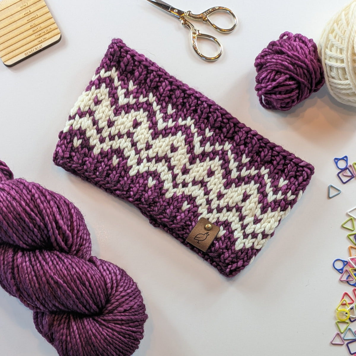 A hand knit fair isle ear warmer headband in cream and plum wine colored yarn, styled in a flat-lay with knitting supplies on a white background.