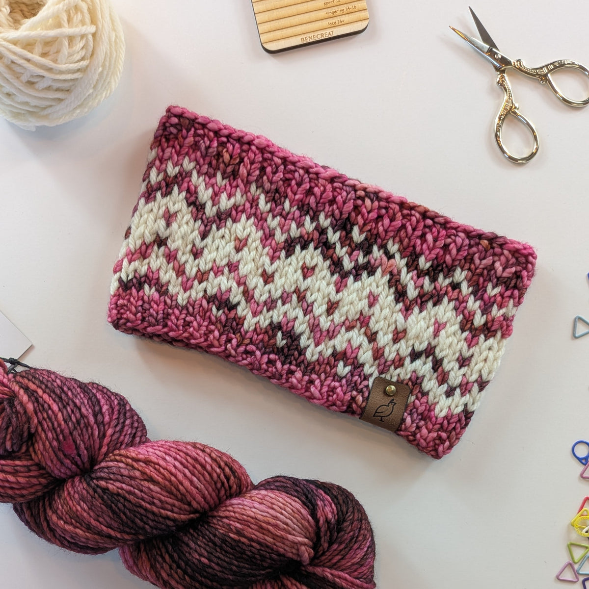 A hand knit fair isle ear warmer headband in a variegated pattern of cream with red, pink, and burgundy yarn, styled in a flat-lay with knitting supplies on a white background.