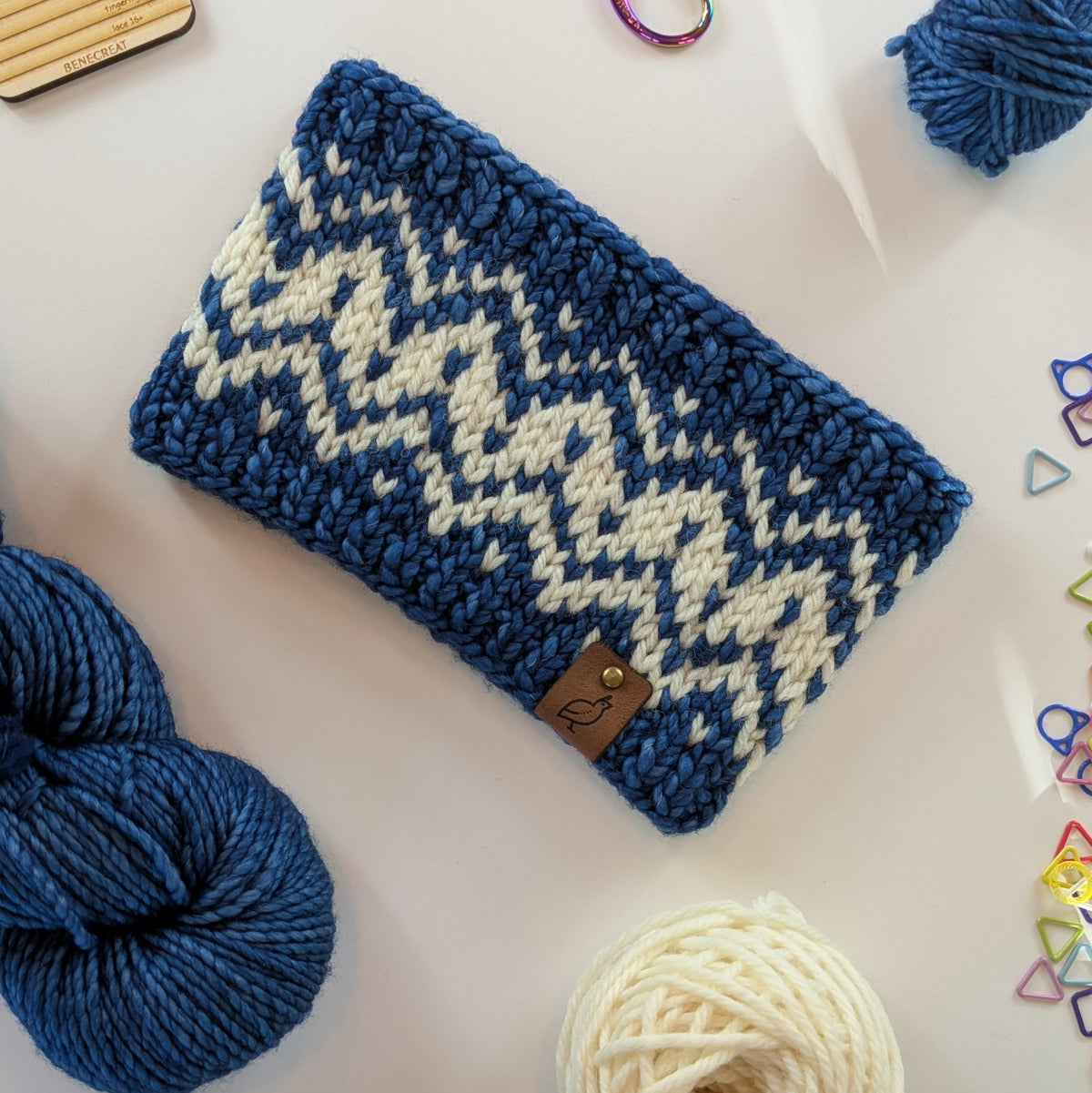 A hand knit fair isle ear warmer headband in cream and blue yarn, styled in a flat-lay with knitting supplies on a white background.