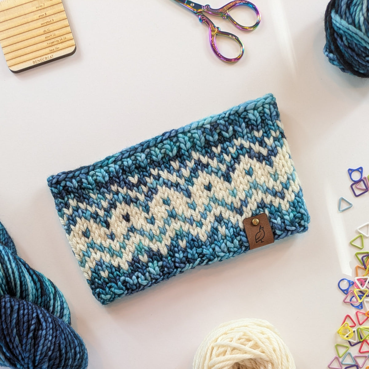 A hand knit fair isle ear warmer headband in a variegated pattern of cream with blue, teal, and aqua colored yarn, styled in a flat-lay with knitting supplies on a white background.