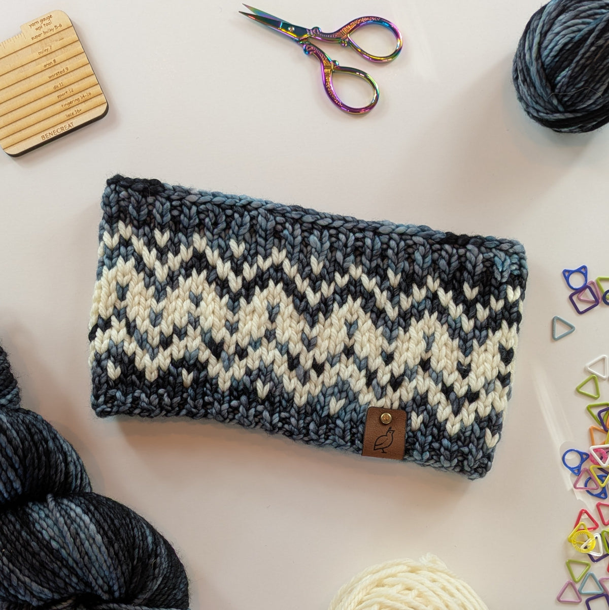 A hand knit fair isle ear warmer headband in a variegated pattern of cream, blue, gray, and black yarn, styled in a flat-lay with knitting supplies on a white background.