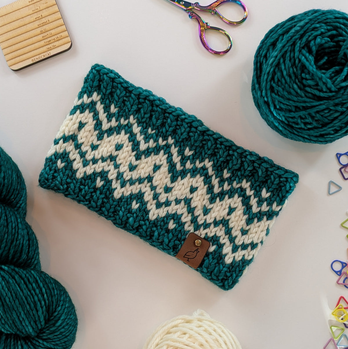 A hand knit fair isle ear warmer headband in cream and teal yarn, styled in a flat-lay with knitting supplies on a white background.