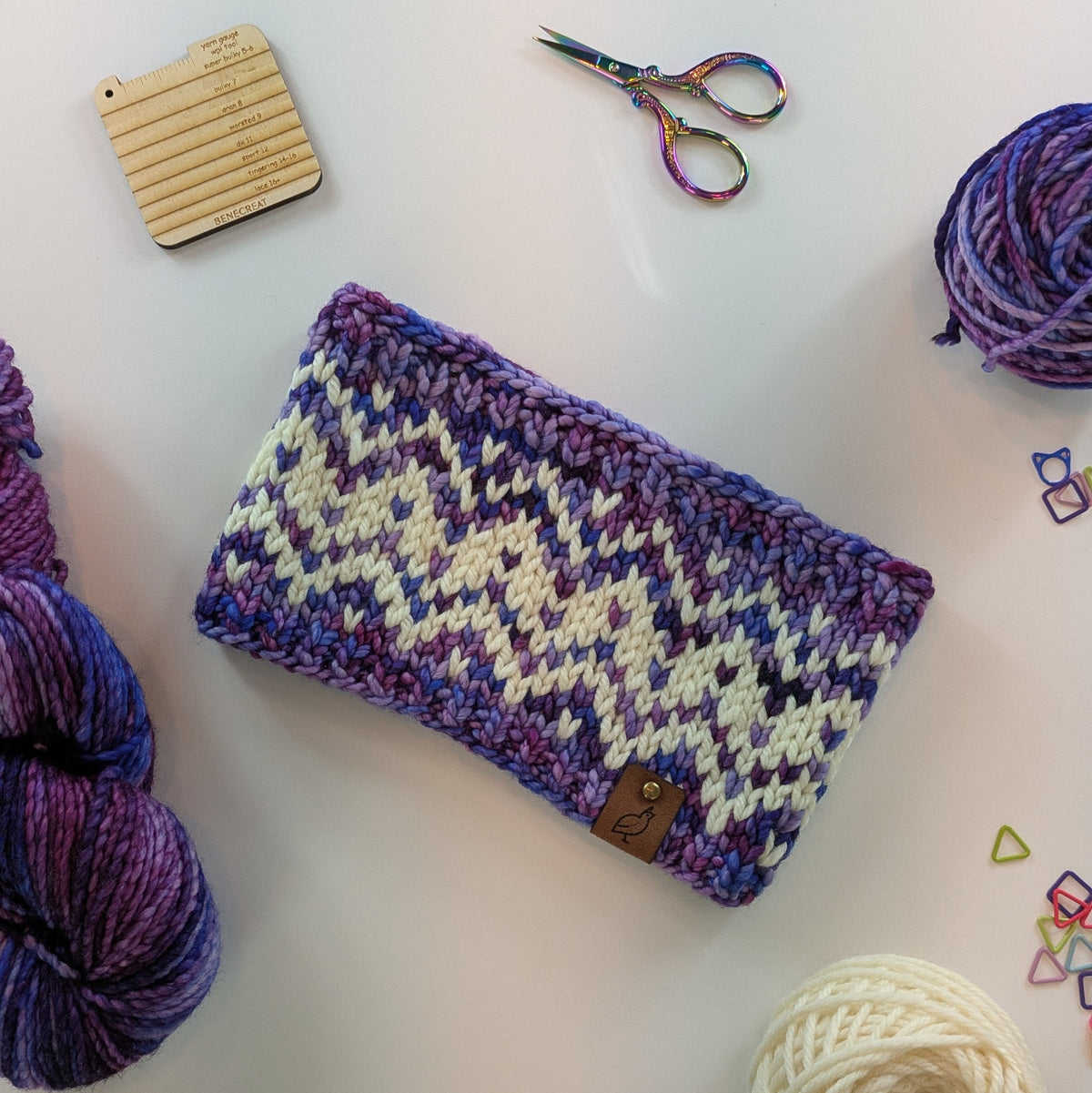 A hand knit fair isle ear warmer headband in a variegated pattern of cream with plum, purple, and blue yarn, styled in a flat-lay with knitting supplies on a white background.